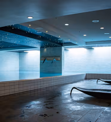 Inomhuspoolområde med modern design, med reflekterande tak och kaklade väggar. Två solstolar finns på det våta stengolvet bredvid poolen. Mjuk belysning skapar en lugn atmosfär. Säkerhetsskyltar är synliga på en kolumn.