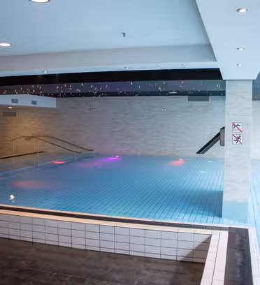 Inomhuspool med ljusblå kakel, omgiven av beiga väggar. Det finns räcken som leder ut i vattnet och levande färgade ljus som reflekteras på ytan. Taket har infälld belysning, vilket skapar en modern atmosfär.