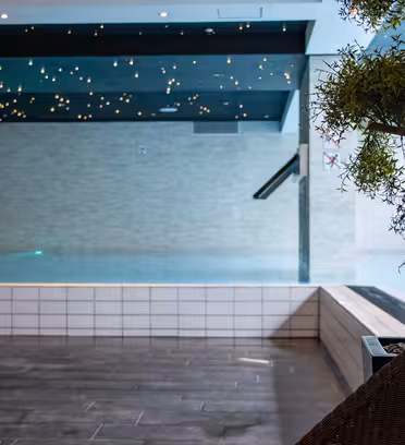 Inomhuspoolområde med en lugn atmosfär, med mjuk belysning och stjärnliknande taklampor. Poolen kantas av vitt kakel, med en krukväxt och en trästol i förgrunden.