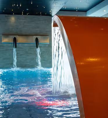 Inomhuspool med böjt orange vatten har vatten som rinner ner i poolen. Ytterligare två väggmonterade piper häller vatten i bakgrunden. Lokalen har en modern design med stenväggar och mörkt tak.