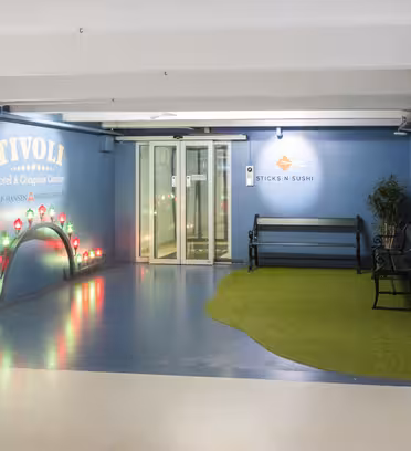 Entréhall med blå väggar, med skylten Tivoli Hotel & Congress Center och Sticks 'N' Sushi-skylten. Det finns en bänk på grönt konstgräs bredvid en glasdörr. Inredningen inkluderar upplyst båge med färgglada lampor till vänster.