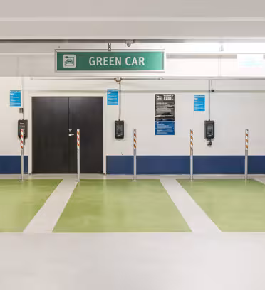 En parkeringsplats med fyra gröna billaddningsplatser markerade med grön färg. Varje utrymme har en vertikal stolpe och en laddare monterad på väggen med en skylt ovanför som läser "GRÖN BIL." Golv och väggar är övervägande vita.