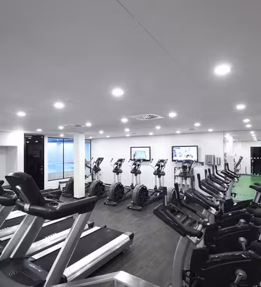 Ett modernt gym med rader av löpband och stationära cyklar. Lokalen är väl upplyst med infälld belysning. Två stora skärmar är monterade på väggen och en glasvägg skiljer området från ett annat rum.