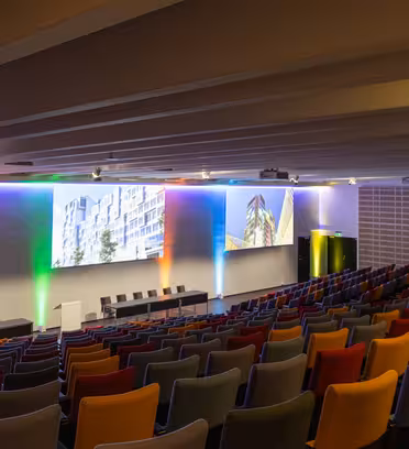 En tom, färgglad auditorium med rader av röda, blåa och gråa stolar mot en scen. Scenen har ett vitt podium och fem svarta stolar. Skärmarna ovan visar arkitektoniska bilder. En projektion av regnbågsljus pryder väggen.