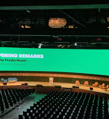 En stor auditorium med rader av tomma platser. En ljusgrön skärm visar texten "Inledande kommentarer" och "Mette Frederiksen, Danmarks premiärminister." Scenen är upplagd med stolar och bord för en presentation.