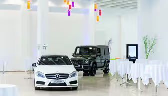 Ett rymligt showroom med en vit Mercedes-Benz A-Klasse-bil i förgrunden och en mörk Mercedes-SUV bakom sig. Området är dekorerat med färgglada hänglampor och flera runda bord täckta med vita dukar.