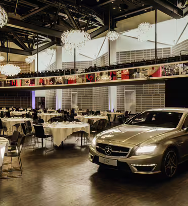 En elegant silverbil står parkerad i en rymlig, elegant hall med trägolv. Lokalen är inredd för ett event, med runda bord täckta av vita dukar, omgivna av svarta stolar. Moderna ljuskronor hänger i taket och ger ett varmt sken.