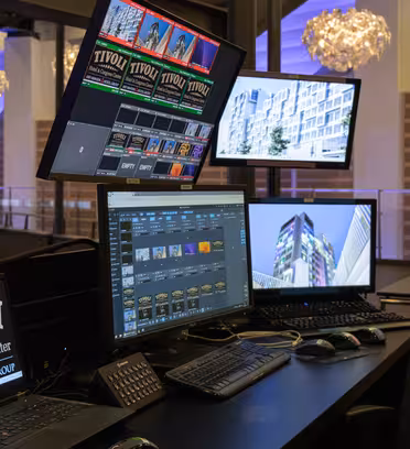 En kontrollrumsinställning med flera monitorer som visar bilder och information. En bildskärm visar affischer för "Tivoli", en annan visar en byggnads exteriör. En bärbar dator med logotypen "Tivoli Hotel & Congress Center" finns till vänster. Ljuskronor hänger ovanför.