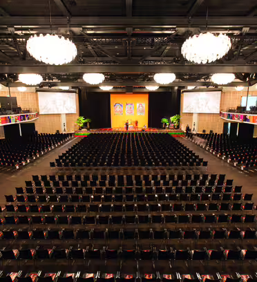 En stor, elegant inredd auditorium med rader av tomma stolar mot en scen prydd med livfulla banderoller. Två skärmar flankerar scenen. Ljuskronor hänger i taket och balkonger kantar sidorna av rummet.