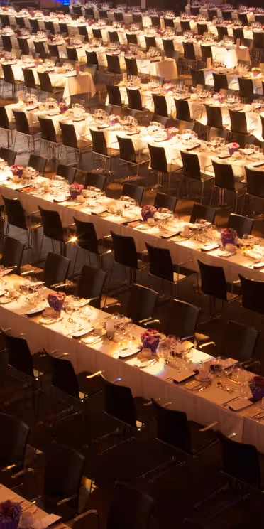 En stor festsal är fylld med rader av långa, elegant dukade bord. Varje bord är prydd med vita dukar, arrangerade tallrikar, bestick och mittstycken med lila blommor. Tomma stolar är snyggt inriktade på båda sidor om borden.