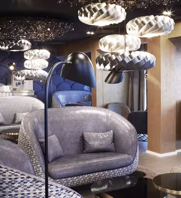 Modern loungedel med snygga, svängda grå soffor och matchande fåtöljer. Unika hängande lampor hänger i taket och ger ett mjukt ljus. Väggarna har en geometrisk design och det finns bord med reflekterande ytor utspridda.