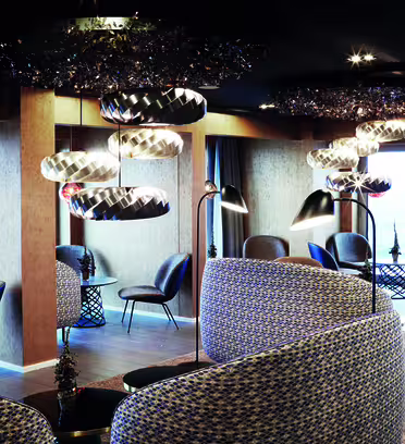 Modern lounge med mjuk belysning, mönstrade soffor och snygga stolar runt små runda bord. Unika, reflekterande armaturer hänger i taket. Stora fönster släpper in naturligt ljus och skapar en mysig men modern atmosfär.